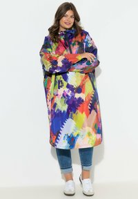 Impermeabile oversize multicolore con cappuccio. Presenta motivi astratti in tonalità vivaci, realizzato in materiale impermeabile. Modello con jeans casual.