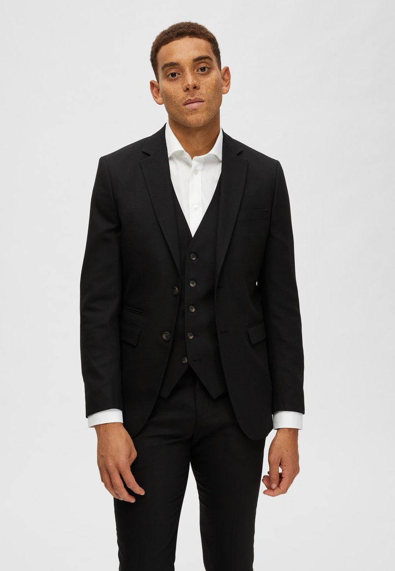 Schwarzes dreiteiliges Anzug mit tailliertem Blazer, Weste mit Knöpfen und schmal geschnittenen Hosen. Knackig weißes Hemd mit französischen Manschetten. Einheitlicher Hintergrund.