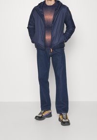 Giacca puffer blu navy con cappuccio, maglione con gradiente blu testurizzato, jeans dritti blu scuro e sneakers nere con accenti gialli e viola.