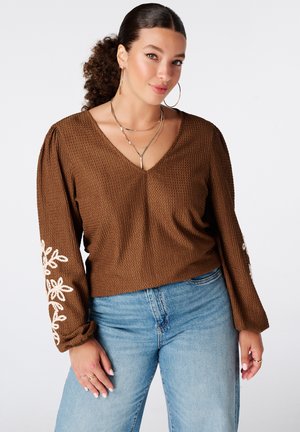 Vrouw met krullend donker haar in een bruin gestructureerd V-hals top met witte bloemenborduursel op de mouwen en lichtblauwe spijkerbroek, met gouden sieraden.