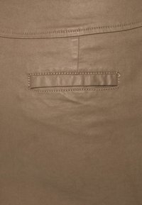 Gros plan d'un tissu beige avec une poche passepoil horizontale et des coutures visibles au-dessus de la poche sur un vêtement.