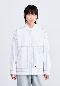 Fehér zipzáras dzseki kontrasztvarrással, álló gallérral és elülső zsebekkel. Nike logó a bal oldalon. Puha anyag, oversized szabás.