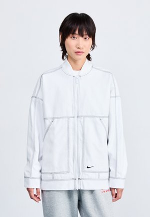 Jachetă albă cu fermoar, cusături contrastante, guler înalt și buzunare frontale. Logo-ul Nike pe partea stângă. Material moale, croială oversized.
