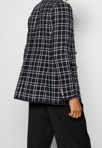 Marinblå kavaj med vitt rutmönster, texturerad tyg och guldkknappar på ärmarna. Skräddarsydd passform med en något utsvängd silhuett.