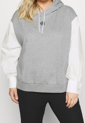 Femme portant un sweat à capuche gris Nike avec des manches longues blanches bouffantes et un pantalon noir, debout devant un fond uni.