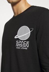 Svart långärmad t-shirt med ett vitt motiv av en planet med en ring, kompletterat med texten "SPACE MSSN NGC 2346" under designen.