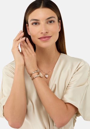 Bracelets en or rose avec une finition lisse, dotés d'un pendentif rond et d'un pendentif en forme de cœur. Le modèle porte un haut en satin beige clair.