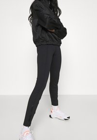 Person som bär svarta dragkedjeleggings, svart jacka, vita strumpor och vita sneakers med orange accent, stående mot en vanlig vit vägg.