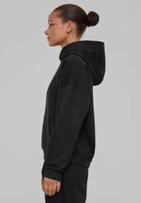 Mujer con el cabello oscuro recogido en un moño, vestido con una sudadera negra holgada con capucha y pantalones a juego, de perfil sobre un fondo gris liso.