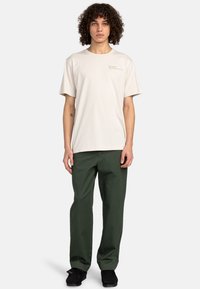 Camiseta beige clara de manga corta con logo verde, combinada con pantalones verde oscuro. Llevando zapatos negros, el modelo se encuentra frente a un fondo plano.