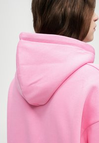 Kapuzenpulover u svijetloružičastoj mješavini pamuka, s zaobljenom kapuljačom i glatkom teksturom, prikazan sa stražnje strane s naglaskom na dio oko vrata.