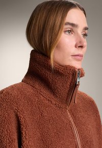 Brown fleece jacket with a high collar, featuring a front zipper and a textured, plush surface. The design is simple and functional.