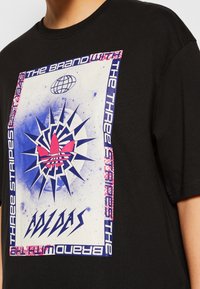 Czarna bawełniana koszulka z dużym wielokolorowym nadrukiem, na którym znajduje się logo Adidas, wyraźne wzory oraz wyraziste elementy tekstowe.