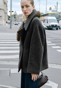 Cappotto grigio in misto lana con ampi rever e accentuato in pelliccia, abbinato a jeans scuri, in piedi su un passaggio pedonale in un contesto urbano.