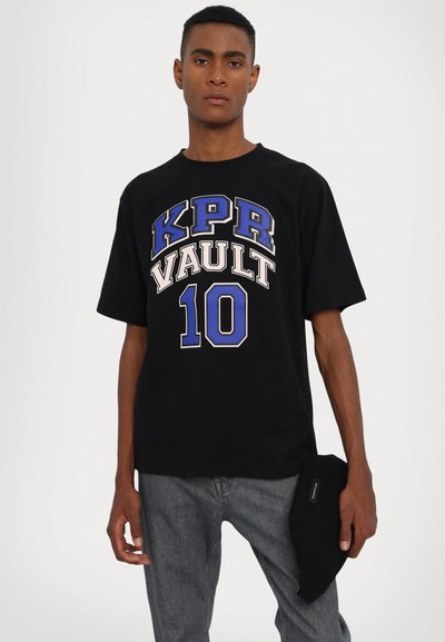 Schwarzes Baumwoll-T-Shirt mit auffälligem "KPR VAULT 10"-Grafik in Blau und Beige. Kurze Ärmel, lockerer Schnitt, kombiniert mit grauen Denim-Hosen und einem schwarzen Accessoire.