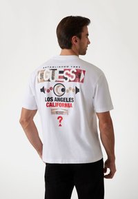 Camiseta de algodón blanca con un estampado gráfico colorido en la parte posterior, que incluye texto y símbolos en rojo, negro y dorado. Mangas cortas.