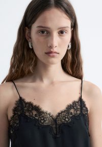 Schwarzes Trägershirt mit dünnen Trägern, das eine Spitze entlang des Ausschnitts aufweist. Glattes Material mit aufwendigen Spitzenverzierungen oben.