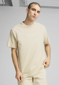 Beige bomulds t-shirt med korte ærmer, prydet med et lyseblåt grafisk design på brystet. Modellen står foran en neutral baggrund.