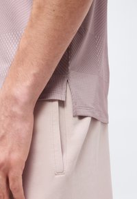 Κοντινό πλάνο του μπράτσου ενός ατόμου δίπλα στην τσέπη μπεζ παντελονιού και πουκάμισου μωβ με γεωμετρικά μοτίβα και πλαϊνή σχισμή στο τελείωμα.