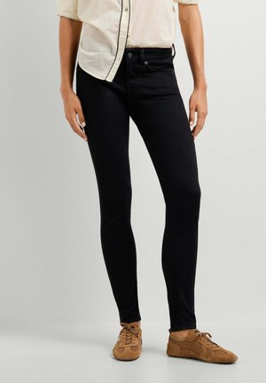 Persona che indossa pantaloni neri slim fit, una camicia crema con bottoni e scarpe casual marroni con lacci, in piedi su uno sfondo uniforme.