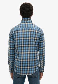 Camicia di flanella a quadri blu con accenti bianchi e neri, caratterizzata da maniche lunghe, colletto button-down e orlo arrotondato. Vista posteriore mostrata.