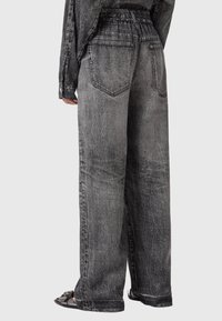 Mujer que lleva unos jeans de mezclilla negros desgastados, de talle alto y corte holgado, con bolsillos traseros y una cinturilla elástica, combinados con sandalias negras.