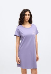 Γυναίκα με σγουρά μαλλιά που φοράει ένα μωβ φόρεμα με κοντά μανίκια και μικρό σχέδιο φοίνικα στο αριστερό στήθος, στέκεται μπροστά σε λευκό φόντο.