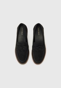 Mocasines de ante negro con detalle trenzado en la parte delantera, suela marrón cosida y logo en el interior. Diseño plano, textura suave.