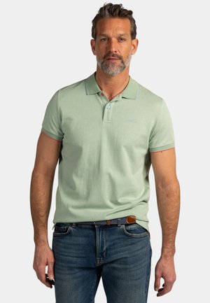 Man met grijze baard, gekleed in een lichtgroen poloshirt met korte mouwen en een blauwe spijkerbroek met een riem met patroon, staand tegen een effen achtergrond.