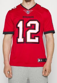 Punainen NFL-pelipaita, jossa joukkueen nimi "Buccaneers", pelaajan numero "12" valkoisena oransseilla yksityiskohdilla, hihoissa musta reuna ja edessä Nike-logo.