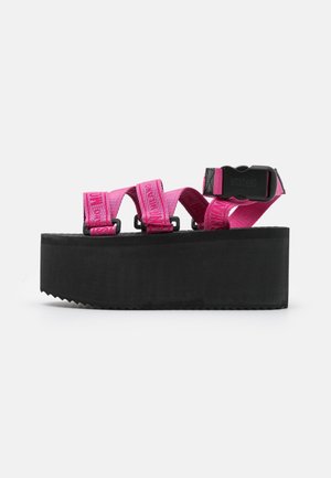 Sandale à plateforme noire avec semelle épaisse et brides réglables rose vif avec boucles en plastique sur fond blanc.