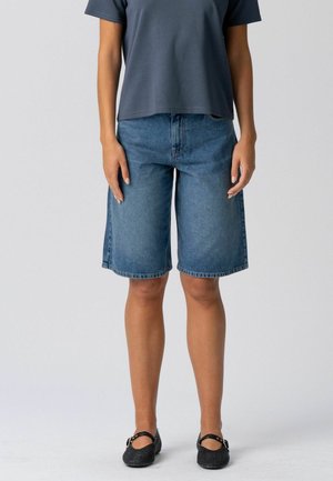 Személy térd alá érő kék farmer rövidnadrágot, sötétszürke pólót és fekete Mary Jane cipőt visel, egyszínű háttér előtt áll.