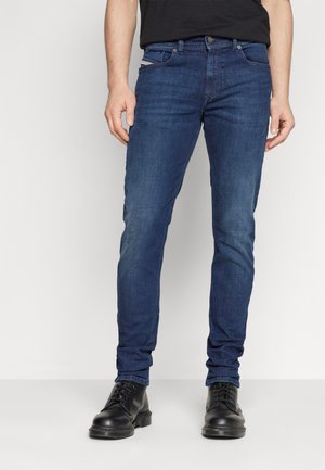 Mann trägt eng anliegende blaue Jeans und schwarze Schnürstiefel, steht vor einem schlichten hellen Hintergrund, Hände entspannt an den Seiten.