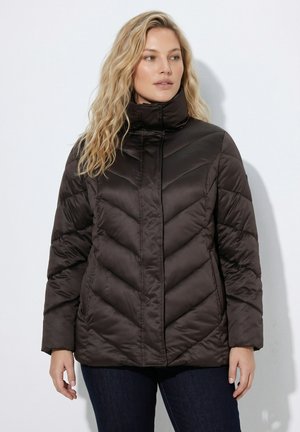 Kvinde med langt blond hår iført en sort dynejakke med høj krave og blå jeans, stående mod en ensfarvet lys baggrund.