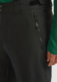 Zwarte outdoorbroek van flexibel materiaal, met een zijzak met rits en een gladde textuur. Taille vastgemaakt met knopen en klittenband.
