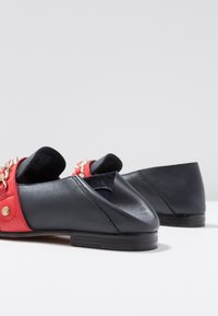 Flats en cuir noir avec une bride rouge ornée de ferrures dorées. Conception à bout pointu, talon plat, et côtés ajourés pour un look ouvert.