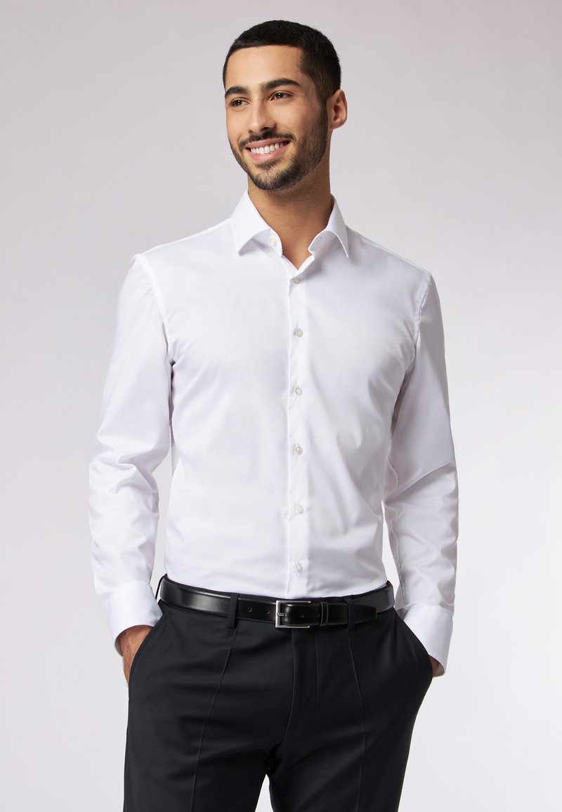 Weißes Langarmhemd mit Button-Down-Kragen, ausgestattet mit Vorderknöpfen und einer glatten Textur. Kombiniert mit schwarzen Hosen und einem Gürtel.