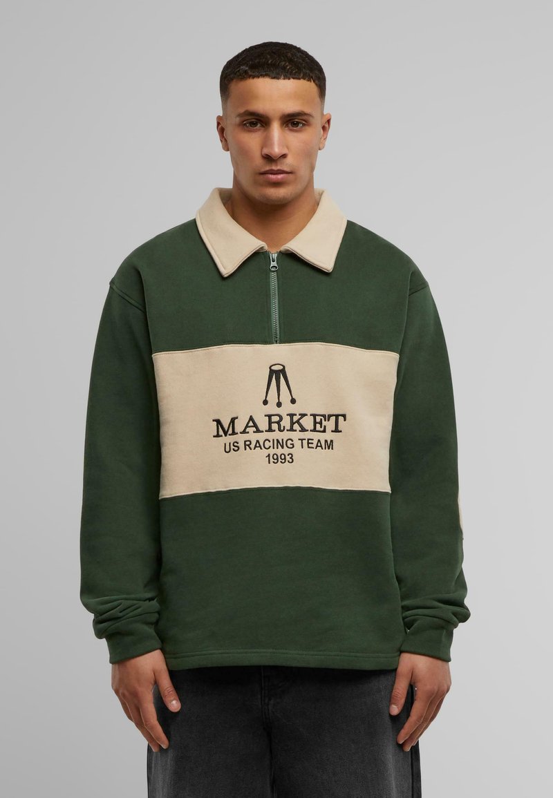 Žalias ir kreminis megztinis su užtrauktuku prie apykaklės, su priekinėmis logotipo spaudėmis "MARKET US RACING TEAM 1993." Minkšta tekstūra.