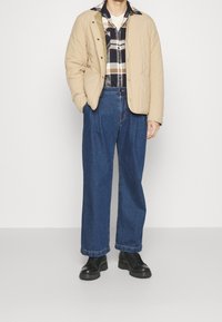 Beige gesteppte Jacke über einem karierten Flanellhemd, kombiniert mit lockeren blauen Jeans und schwarzen Schuhen.