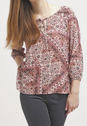 Žena v růžovo-bílé květované blůze s tříčtvrtečními rukávy a tmavě šedých kalhotách pózující s jednou rukou na boku.