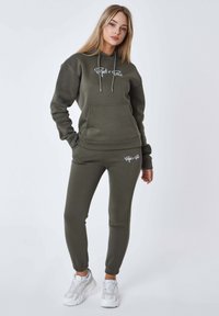 Completo con felpa e pantaloni sportivi color verde oliva in tessuto morbido, dotato di tasca frontale e stampa bianca del logo. Polsini e vita elasticizzati per un maggiore comfort.