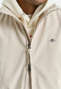 Lichtbeige jas met een rits aan de voorkant, met een klein logo op de borst en een gewatteerde witte sweatshirt met capuchon eronder.