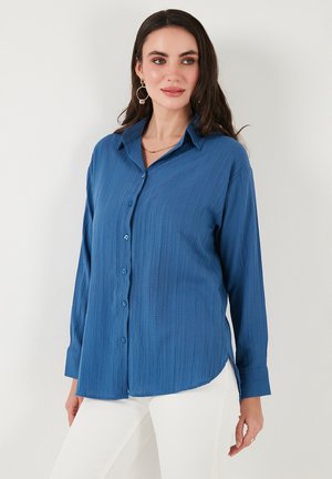Blue long-sleeve shirt with a textured fabric, button-front design, classic collar, and curved hem, paired with white pants.