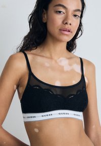 Crni bralette s čipkastim dizajnom, prozirnim mrežastim slojem i širokim bijelim remenom s markom "GUESS". Tanke podesive naramenice.
