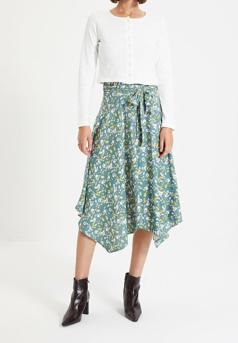 Femme portant un haut blanc à manches longues boutonné, une jupe midi verte à fleurs avec ceinture nouée, et des bottines noires à talons, debout sur fond blanc.