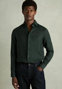 Cămașă din in de culoare verde închis, cu mâneci lungi și design cu nasturi, având guler și un finisaj texturat subtil. Asortată cu blugi de culoare albastru închis.