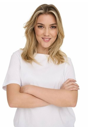 Donna bionda con i capelli sciolti sorride con sicurezza, braccia incrociate, indossa una maglietta bianca semplice a maniche corte su sfondo bianco.