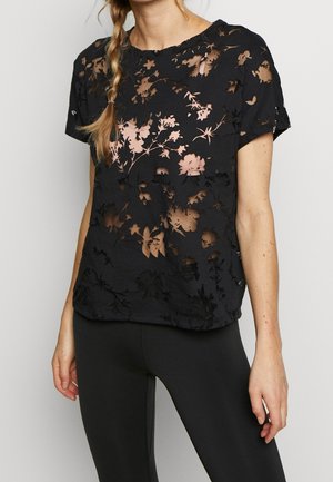 Žena v černém krátkém topu s průsvitnými květinovými výřezy na odhaleném trupu a černých legínách, stojící před jednobarevným pozadím.