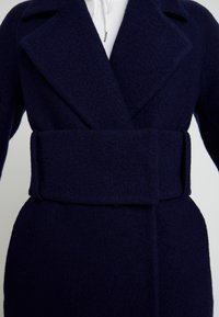 Marineblauer Wollmischmantel mit strukturierter Oberfläche, großen Revers und einem breiten Gürtel in der Taille; zeichnet sich durch eine taillierte Silhouette aus.