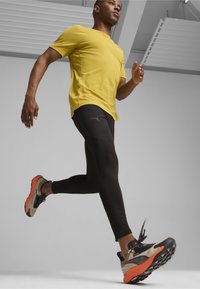 Maglietta sportiva gialla a maniche corte, leggings neri e scarpe da corsa con dettagli arancioni. Il corridore è in movimento su uno sfondo neutro.